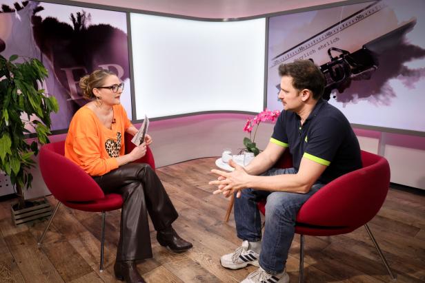 Zwei Personen sitzen sich in roten Sesseln gegenüber und führen ein Gespräch in einem modern eingerichteten Studio.