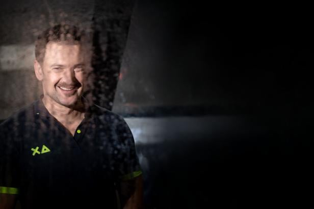 Ein Mann mit dunklem Polohemd lächelt, während er hinter einer nassen, teils beschlagenen Glasscheibe steht.