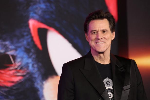 Ein Mann im schwarzen Anzug lächelt vor einem bunten, unscharfen Hintergrund mit roten und blauen Akzenten.