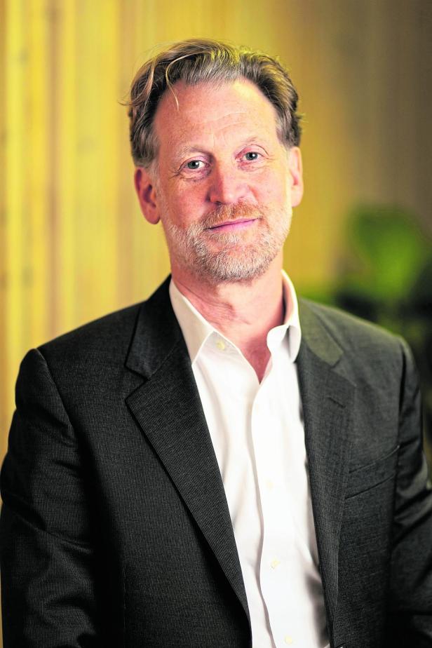 Ein Mann mit grauem Bart und Anzugjacke steht vor einem unscharfen, gelblichen Hintergrund.