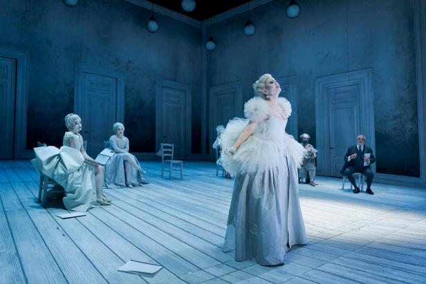 Szenenbild aus "L'opera seria" im Theater an der Wien: Mehrere Personen in barocken Kostümen sitzen und stehen in einem spärlich möblierten, blau beleuchteten Raum mit Holzboden.