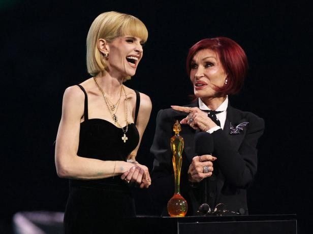 Zwei elegant gekleidete Frauen stehen lachend auf einer Bühne neben einer goldenen Trophäe.