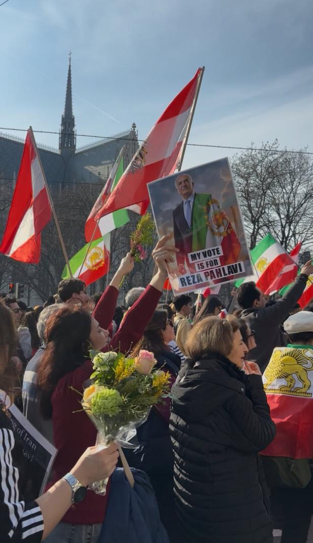 Menschenmenge mit Blumen und Fahnen, ein Plakat mit einer Person und der Aufschrift „My vote is for Reza Pahlavi“ wird hochgehalten.