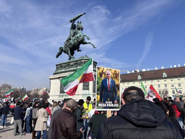 Menschenmenge mit iranischen Flaggen und einem Plakat von Reza Pahlavi vor einer Reiterstatue auf einem Platz.