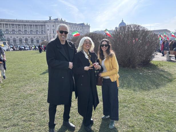 Drei Menschen stehen lachend auf einer Wiese vor einem historischen Gebäude, im Hintergrund sind viele Fahnen zu sehen.