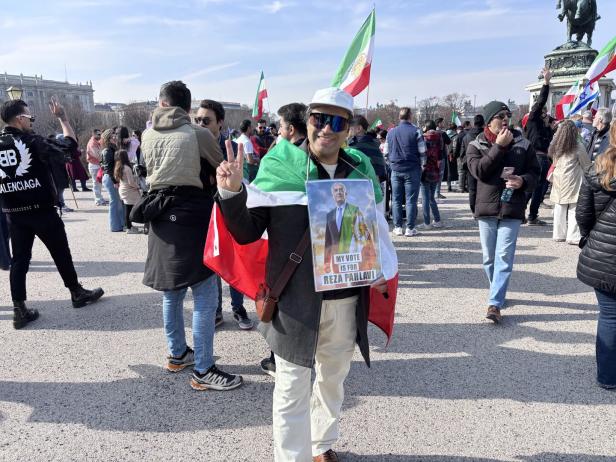 Ein Mann mit einer Flagge und einem Plakat für Reza Pahlavi zeigt das Victory-Zeichen bei einer Kundgebung im Freien.
