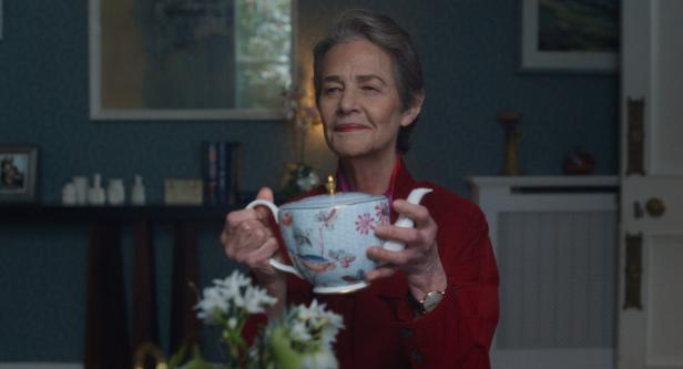 Filmszene: Charlotte Rampling in rotem Blazer hält eine verzierte Teekanne mit Blumenmuster und lächelt leicht.