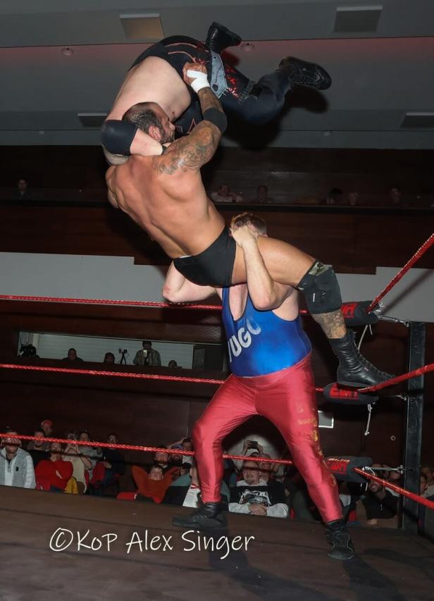 Drei Wrestler führen einen spektakulären Move im Ring vor Publikum aus, einer wird kopfüber hochgehoben.