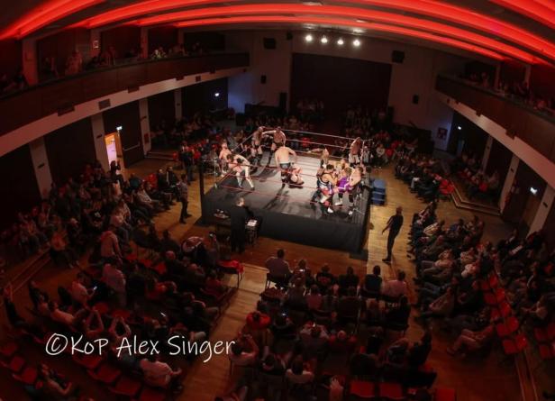 Mehrere Wrestler kämpfen in einem Ring in einer gut besuchten Halle mit rot beleuchteter Decke.
