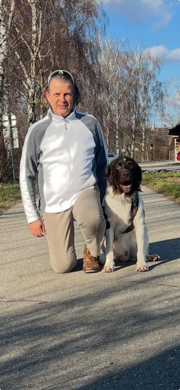 Ein Mann kniet neben einem schwarz-weißen Hund auf einem Weg, im Hintergrund stehen kahle Bäume.
