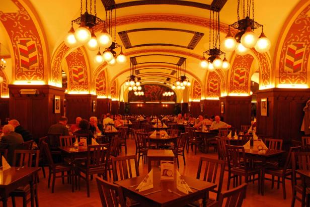 Gäste sitzen in einem traditionsreichen Leipziger Kaffeehaus mit historischer Decke und stimmungsvoller Beleuchtung an Holztischen und genießen Speisen und Getränke.