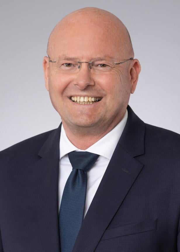 Ein lächelnder Mann mit Brille und Anzug vor grauem Hintergrund.