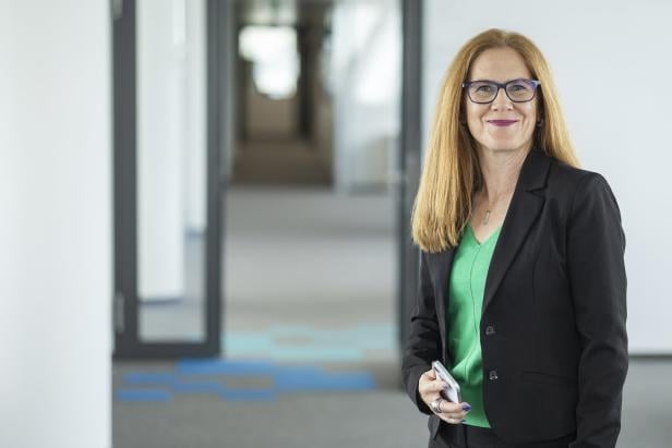 Frau mit langen roten Haaren, Brille und grünem Pullover unter schwarzem Blazer steht lächelnd in einem modernen Büroflur.