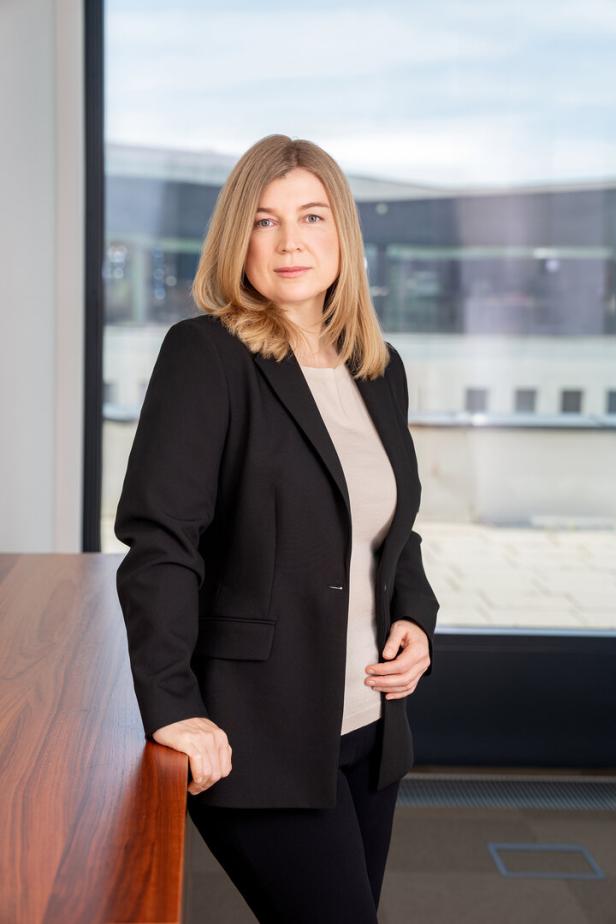 Eine Frau mit blonden Haaren trägt einen schwarzen Blazer vor einem Fenster.