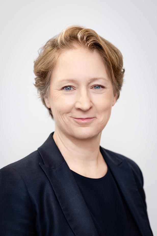 Frau mit kurzen, hellbraunen Haaren und blauem Blazer lächelt freundlich vor hellem Hintergrund.