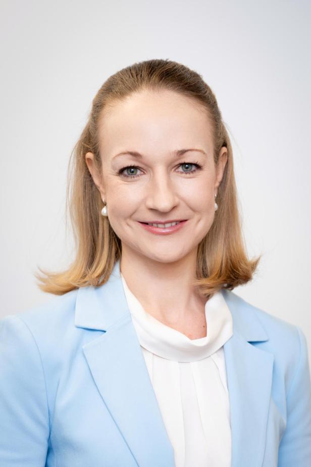 Frau mit schulterlangem blondem Haar, hellblauem Blazer und weißer Bluse lächelt freundlich vor hellem Hintergrund.