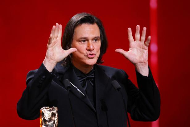 Jim Carrey gestikuliert auf bei den César Awards der Bühne vor rotem Hintergrund.
