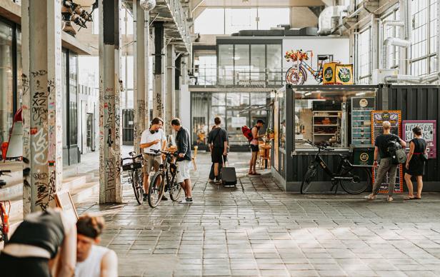 Menschen treffen sich in einer kreativ gestalteten, urbanen Halle der Leipziger Spinnerei, umgeben von Streetart, Fahrrädern und einem modernen Imbissstand.