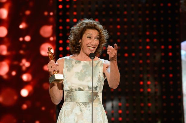 Adele Neuhauser mit lockigem Haar hält eine goldene Statue und spricht in ein Mikrofon.