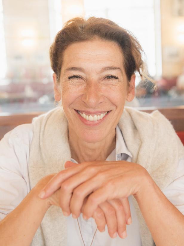 Eine lächelnde Adele Neuhauser mit hellbraunen Haaren sitzt mit verschränkten Händen an einem Tisch.