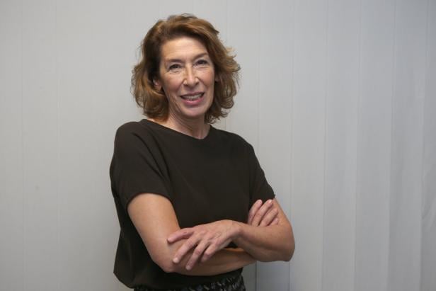 Adele Neuhauser mit braunen Haaren steht mit verschränkten Armen vor einem hellen Hintergrund.