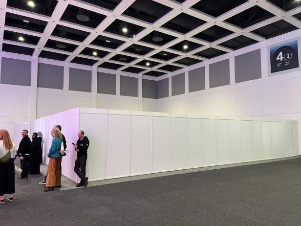 Mehrere Menschen stehen in einer Messehalle vor weißen Trennwänden unter einer hohen Decke mit sichtbaren Lampen.