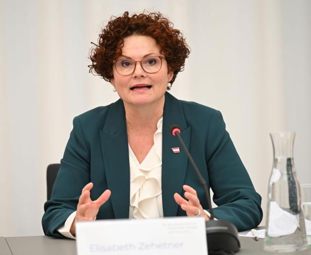 Eine Frau mit lockigen Haaren und Brille spricht an einem Tisch mit Mikrofon und Wasserkaraffe vor sich.