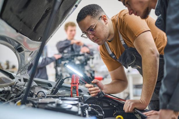 Ein Mechaniker mit Brille untersucht einen offenen Motorraum mit einer Lampe, während andere im Hintergrund arbeiten.