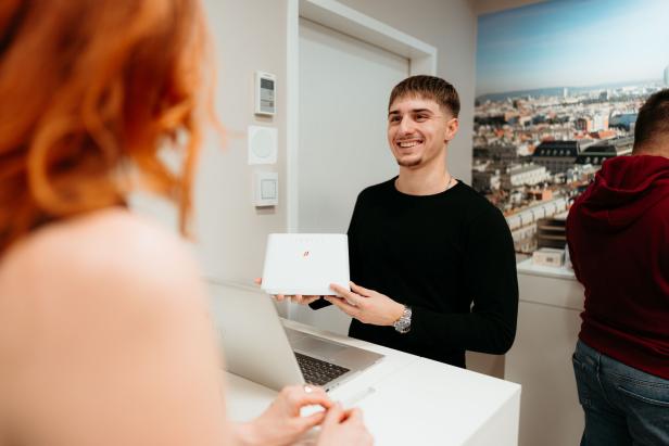 Ein junger Mann hält lächelnd einen weißen Router und spricht mit einer Frau an einem Tresen.