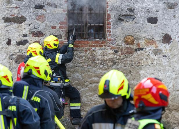 Mehrere Feuerwehrleute mit gelben Helmen stehen vor einer Wand, eine Person führt Löscharbeiten durch ein Fentster durch.