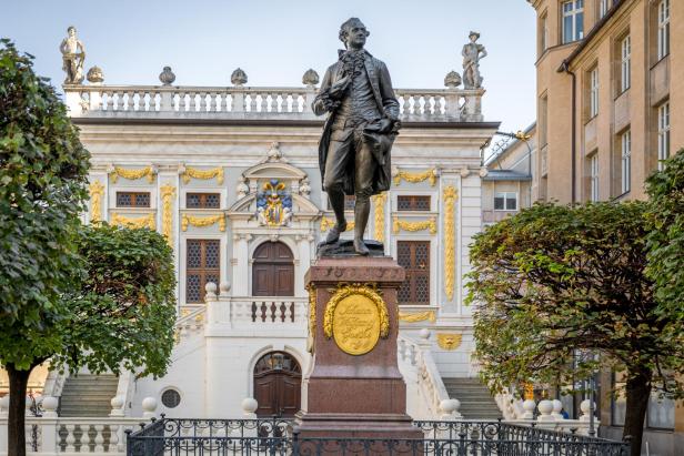 Eine Statue von Johann Wolfgang von Goethe steht vor dem alten Börsengebäude in Leipzig. Das Gebäude hat dekorative gelbe und weiße Züge mit Statuen auf dem Dach. Bäume und ein schwarzer Zaun umgeben die Statue. Eine Treppe führt zum Eingang des Gebäudes hinter der Statue.
