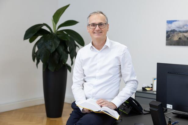 Wolfgang Urbantschitsch sitzt an einem Schreibtisch, hält ein aufgeschlagenes Buch und lächelt.