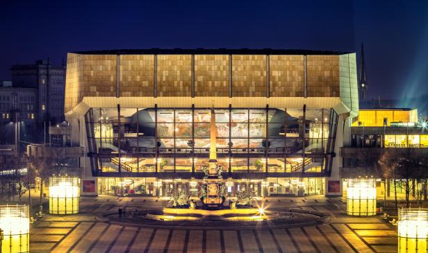 Das Gewandhaus in Leipzig wird bei Nacht von vorne gezeigt. Das große Gebäude hat ein modernes Design mit vielen Fenstern. Helle Lichter beleuchten das Äußere und davor steht ein Springbrunnen. 