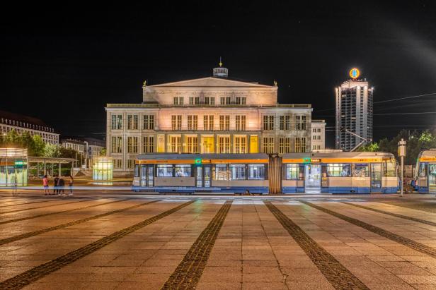 Ein Foto der Oper Leipzig in Leipzig, Sachsen, bei Nacht. Das Gebäude ist beleuchtet und es fährt eine Straßenbahn davor.