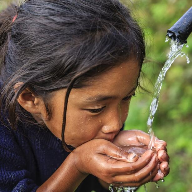 Ein junges Mädchen trinkt Wasser, das aus einem Rohr in ihre Hände fließt.