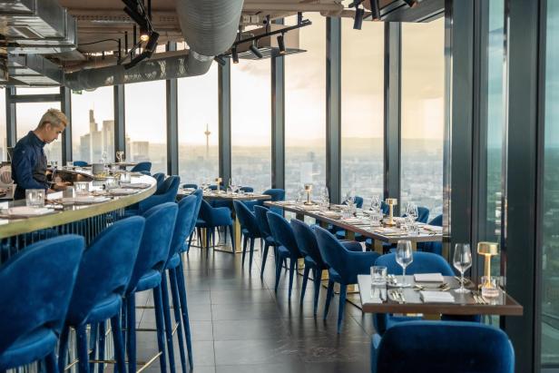 Modernes Restaurant mit blauen Samtstühlen und Panoramablick auf die Skyline einer Großstadt.