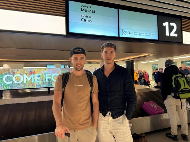 Zwei junge Männer stehen an einem Gepäckband am Flughafen, im Hintergrund warten weitere Reisende auf ihr Gepäck.