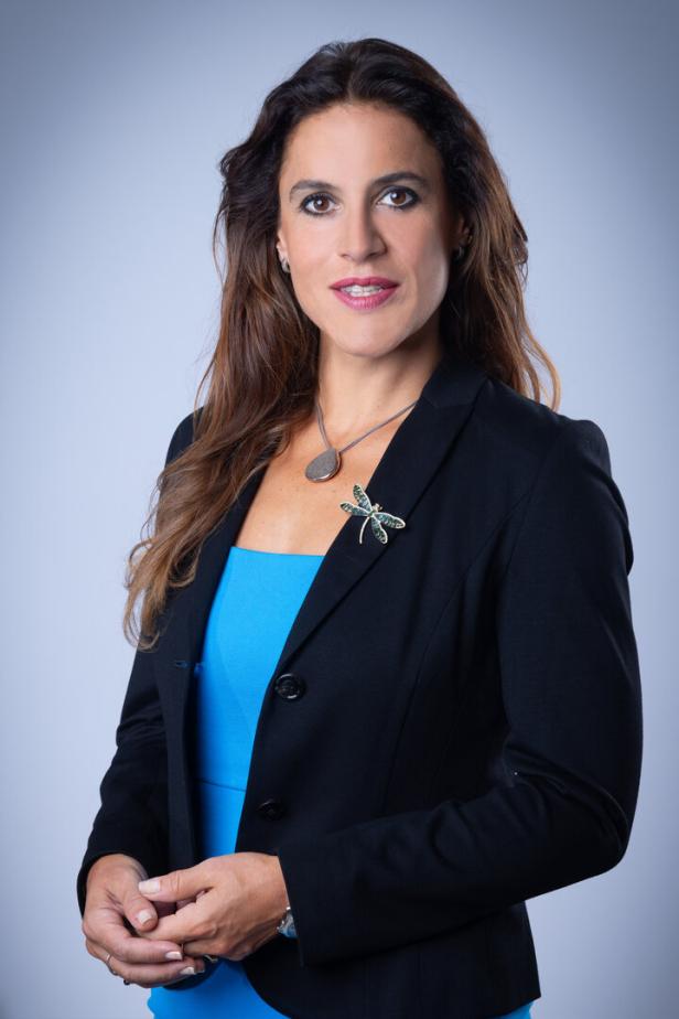 Eine Frau mit braunen Haaren trägt ein blaues Kleid und einen schwarzen Blazer.