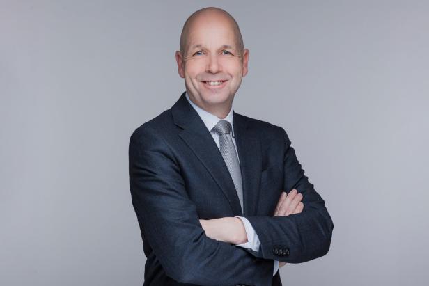 Ein lächelnder Mann im Anzug mit verschränkten Armen vor neutralem Hintergrund.