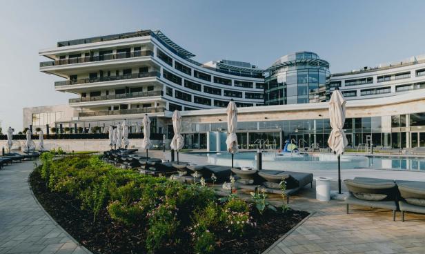 Modernes Hotelgebäude mit Pool, Liegen und geschlossenen Sonnenschirmen im Außenbereich.