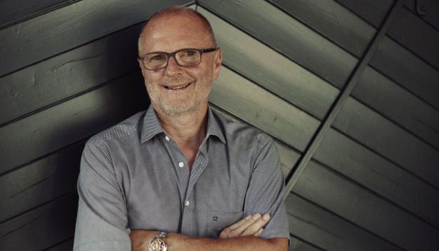 Ein älterer Mann mit Brille und grauem Hemd steht lächelnd mit verschränkten Armen vor einer dunklen Holzvertäfelung.