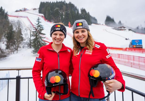 Zwei Frauen in roten Jacken mit Helmen stehen lächelnd vor einer verschneiten Skipiste und tragen Mützen mit Sponsorenlogos.