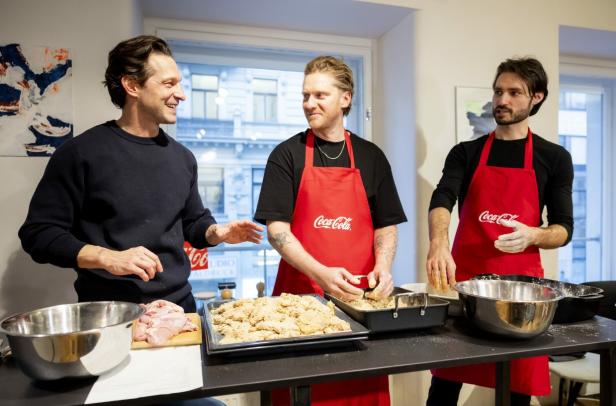 Drei Männer bereiten gemeinsam paniertes Hähnchen in einer Küche mit Coca-Cola-Schürzen zu.