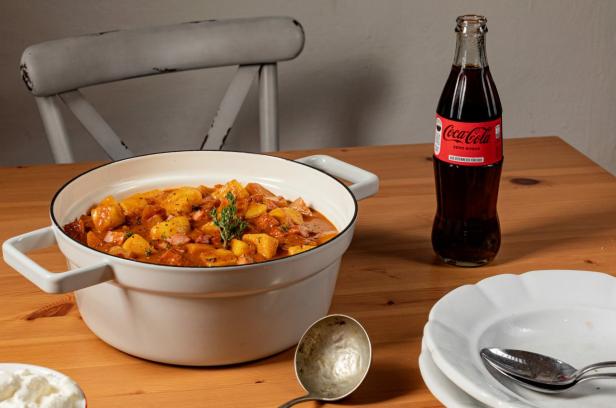 Erdäpfelgulasch in einem weißen Topf neben einer Flasche Coca-Cola Zero auf Holztisch.