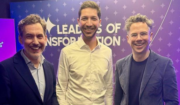 Drei Männer stehen lächelnd vor einer lila Wand mit der Aufschrift „Leaders of Transformation“.