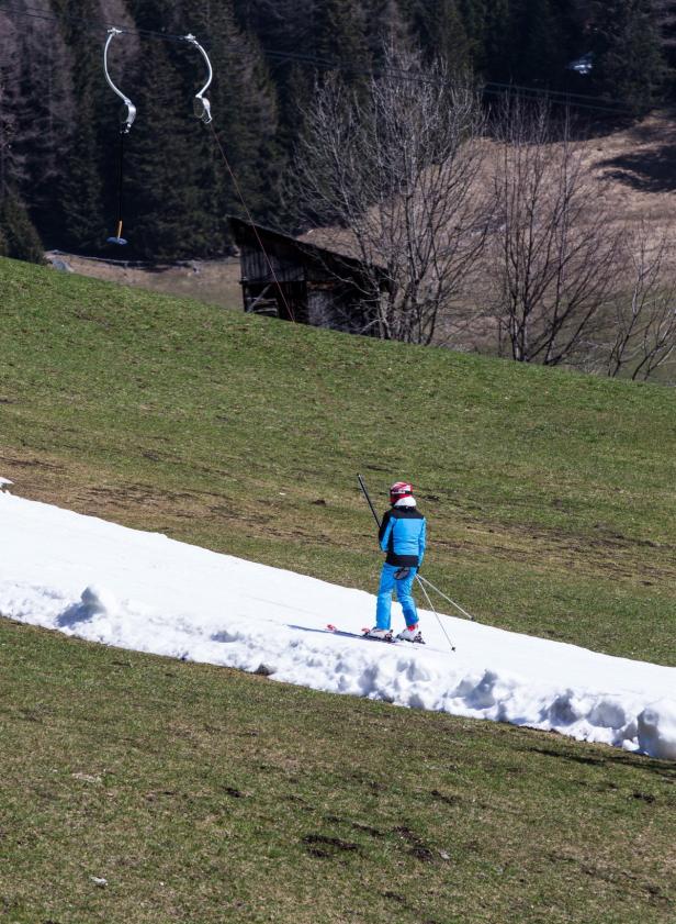 Eine Person fährt Ski auf einem schmalen Schneeband einen grünen Hügel hinauf, ein Schlepplift im Hintergrund.