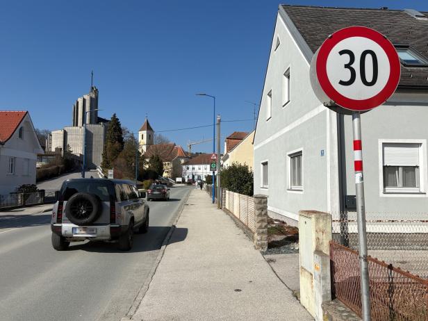 Straße mit Tempo-30-Schild, geparkten Autos, Wohnhäusern und einer Kirche im Hintergrund bei klarem Himmel.