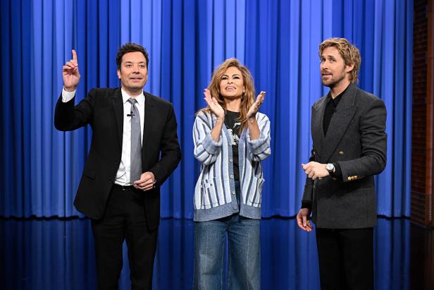 Jimmy Fallon, Eva Mendes und Ryan Gosling auf einer Bühne vor einem blauen Hintergrund. Mendes klatscht.
