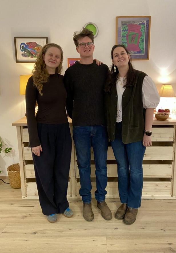 Drei junge Erwachsene stehen lächelnd nebeneinander in einem gemütlich eingerichteten Raum mit Bildern und Lampen an der Wand.