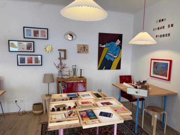 Heller Raum mit Tischen, Kunstmaterialien, roten Stühlen, bunten Bildern an den Wänden und zwei Hängelampen.